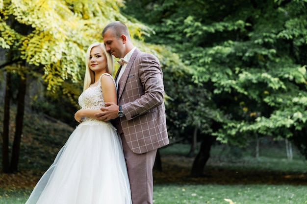 Giovani coppie che godono dei momenti romantici mentre camminano nel parco. Sposa e sposo alla moda che posano e che baciano nel parco il giorno delle nozze. Sposa elegante in bellissimo abito bianco, sposo in un abito.