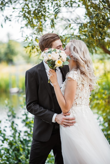 Giovani coppie che godono di momenti romantici mentre si cammina nel parco. Sposa e sposo alla moda che posano e si baciano nel parco il giorno delle nozze. Sposa elegante in bellissimo abito bianco, sposo in abito