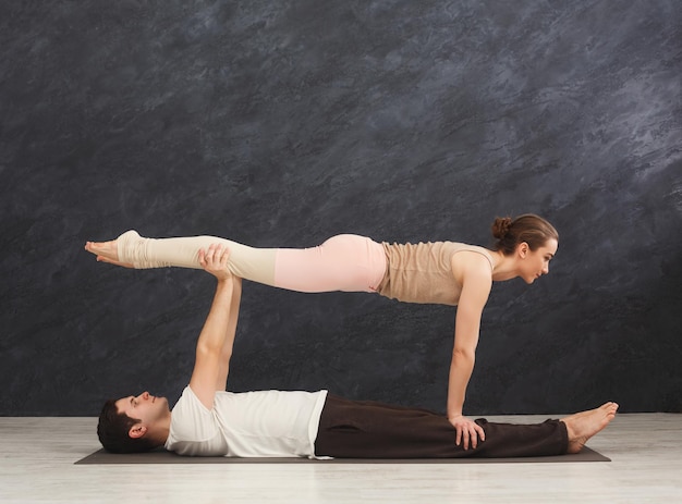 Giovani coppie che praticano insieme acroyoga sulla stuoia in palestra. Donna che fa posa della plancia, copia spazio, vista laterale. Partner yoga, flessibilità, concetto di fiducia