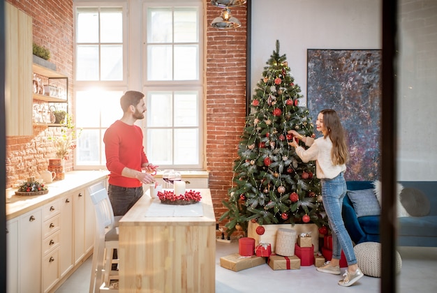 Giovani coppie che si preparano per un nuovo anno decorando il loro bellissimo appartamento in piedi nella sala da pranzo con l'albero di Natale sullo sfondo