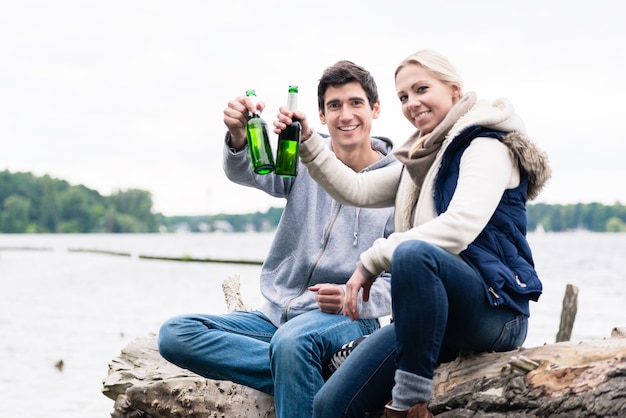 Giovani coppie che si siedono sul ceppo di albero in riva al fiume a bere birra e dire applausi