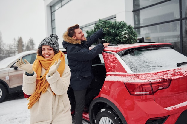 Giovani coppie che stanno vicino all'automobile con l'albero sulla parte superiore. Insieme all'aperto in inverno.