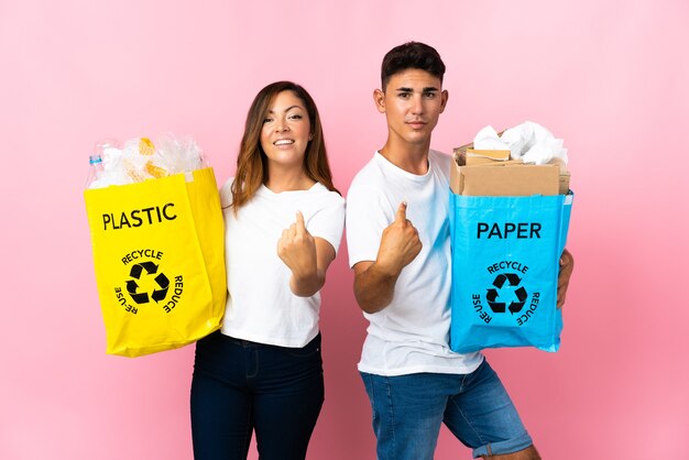 Giovani coppie che tengono un sacchetto pieno di plastica e carta sul colore rosa che fa gesto venente