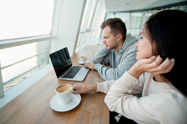 Giovani coppie che trascorrono del tempo al bar e guardano video sul computer portatile. Donna e uomo che bevono caffè e guardano foto insieme. Caffè sullo sfondo. Donna che si aggiusta i capelli quando l'uomo usa il laptop.