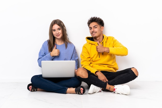 Giovani coppie con un computer portatile che si siede sul pavimento su bianco che dà un pollice in alto gesto
