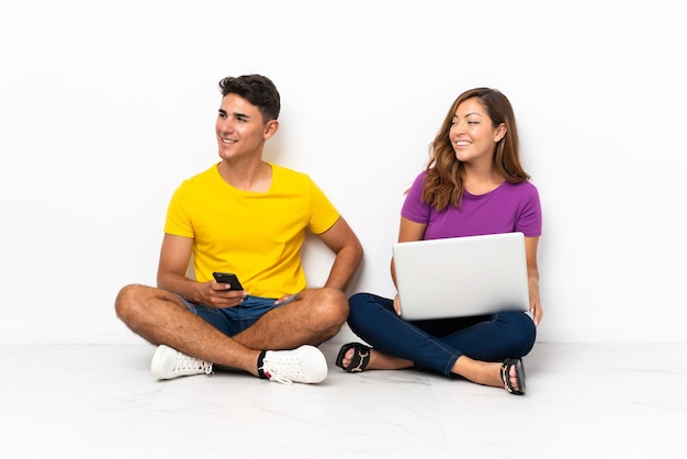 Giovani coppie con un computer portatile che si siede sul pavimento su bianco che osserva al lato e che sorride