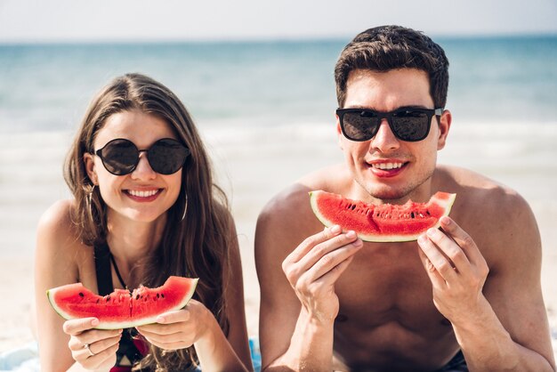 Giovani coppie degli amanti romantici che si rilassano tenuta e che mangiano una fetta di anguria sulla spiaggia tropicale Vacanze estive