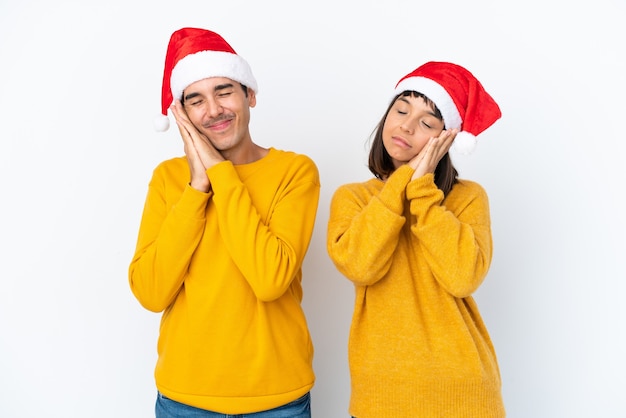 Giovani coppie della corsa mista che celebrano il Natale isolato su fondo bianco che fa il gesto di sonno nell'espressione adorabile