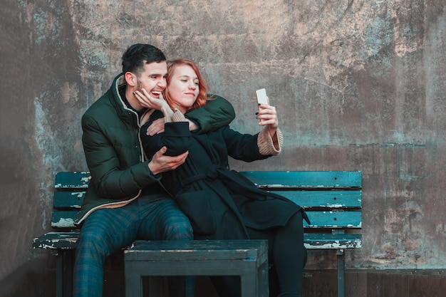 Giovani coppie emotive allegre che si siedono sulla panchina e che fanno Selfie. Due persone felici amano la storia sulla strada