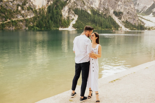 Giovani coppie felici che abbracciano e che sorridono mentre levandosi in piedi vicino al lago di Braies, Italia