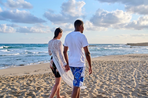 Giovani coppie felici che camminano sulla spiaggia tenendosi per mano indietro vista