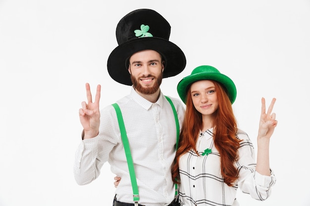 Giovani coppie felici che indossano costumi, celebrando il giorno di StPatrick isolato sopra il muro bianco, divertendosi insieme, gesto di pace