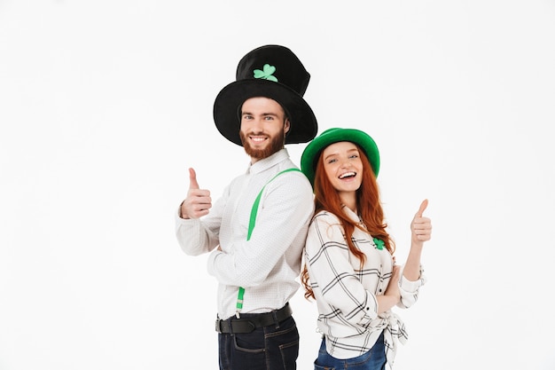 Giovani coppie felici che indossano costumi, celebrando il giorno di StPatrick isolato sopra il muro bianco, divertendosi insieme, pollice in alto