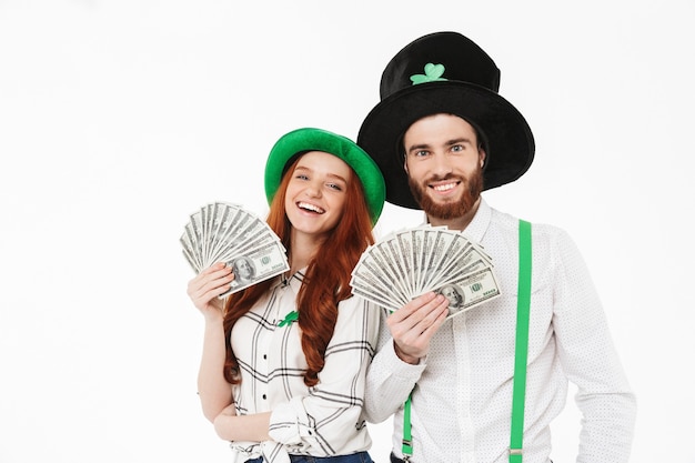 Giovani coppie felici che indossano costumi, celebrando il giorno di StPatrick isolato sopra il muro bianco, tenendo le banconote dei soldi