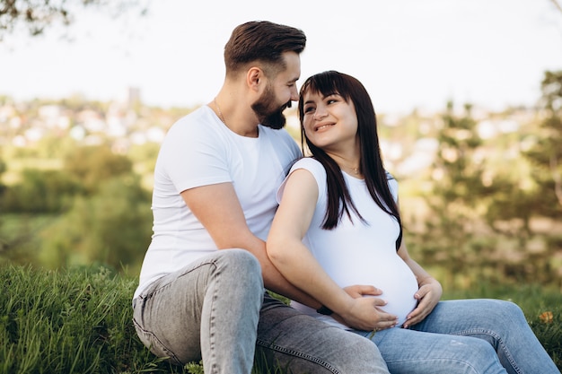 Giovani coppie felici che prevedono bambino, donna incinta con la pancia commovente del marito, sedentesi sull&#39;erba verde