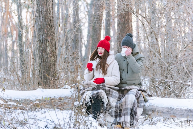 Giovani coppie felici che si abbracciano in un parco invernale innevato avvolto in una coperta e bevendo il tè dalle tazze