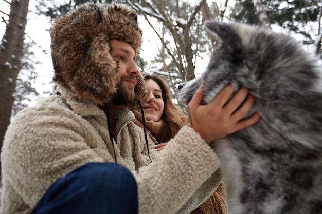 Giovani coppie felici in abbigliamento invernale che giocano con il husky siberiano di razza e si divertono nella pineta innevata