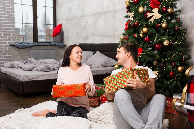 Giovani coppie felici in pigiama che ridono e si rallegrano dei loro regali mentre sono seduti sul tappeto vicino all'albero di Natale nella stanza in stile loft