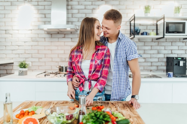 Giovani coppie graziose romantiche che cucinano insieme nella cucina moderna bianca con la verdura e la frutta sullo scrittorio.