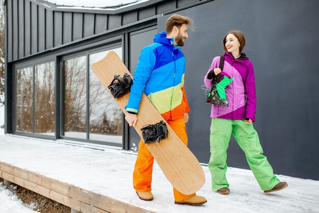 Giovani coppie in tute da sci colorate che camminano insieme allo snowboard vicino alla casa moderna o all'hotel in montagna