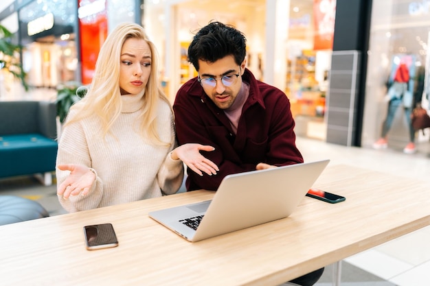 Giovani coppie perplesse che utilizzano insieme il computer portatile di battitura che discutono l'acquisto su Internet che osserva allo schermo che si siede alla tavola nella sala del centro commerciale