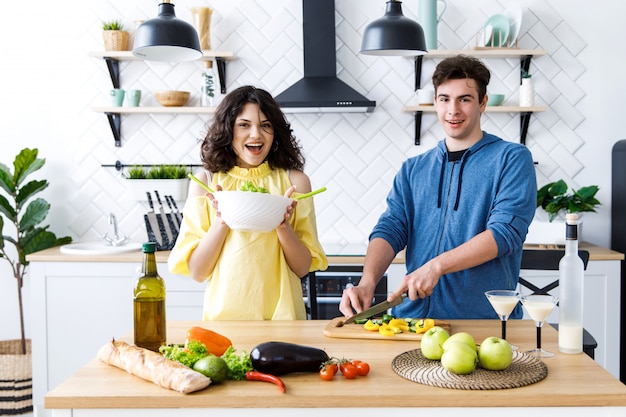 Giovani coppie sorridenti sveglie che cucinano insieme alla cucina a casa. I giovani stanno preparando un'insalata in una buona cucina
