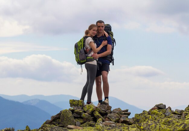 Giovani coppie turistiche con gli zainhi, l'uomo atletico e la ragazza graziosa che stanno abbracciati sulla cima della montagna rocciosa su panorama nebbioso della montagna. Concetto di turismo, viaggi, arrampicata e amicizia.