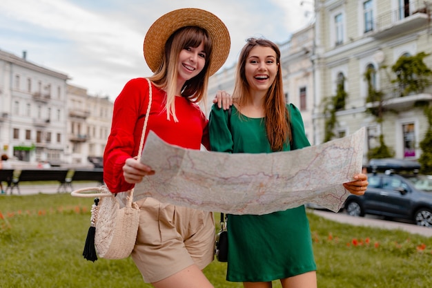 Giovani donne alla moda che viaggiano insieme in Europa vestite con abiti alla moda primaverili e accessori che tengono mappa