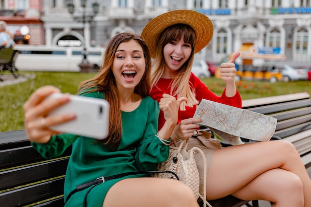 Giovani donne alla moda che viaggiano insieme in Europa vestite in abiti e accessori alla moda primaverili sorridenti amici felici divertendosi a scattare foto sulla mappa della holding della fotocamera del telefono