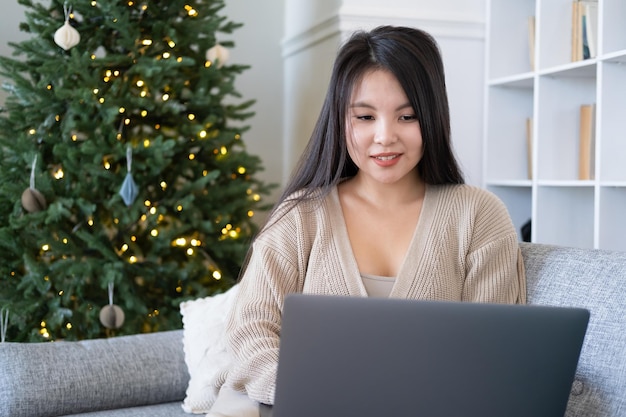 giovani donne asiatiche attraenti in abiti accoglienti utilizzando il laptop sul divano a casa, periodo natalizio