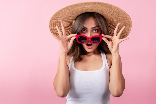 Giovani donne asiatiche turisti che indossano cappelli di paglia a tesa larga e occhiali da sole rossi. Ha espresso sorpresa. Isolato su sfondo rosa