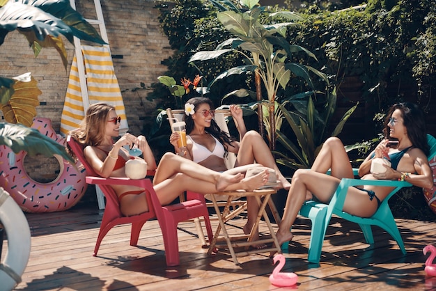 Giovani donne attraenti in costume da bagno che bevono cocktail e sorridono mentre si rilassano a bordo piscina all'aperto