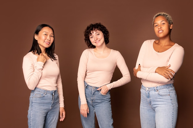 Giovani donne gioiose in pullover bianchi e jeans blu in piedi l'una vicino all'altra su sfondo marrone e ti guardano con sorrisi a trentadue denti