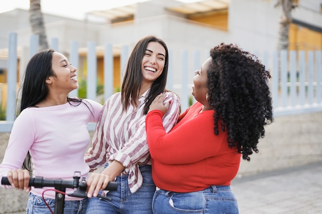 Giovani donne latine alla moda con diversi colori del corpo e della pelle che si divertono in città con il monopattino elettrico - Ragazze multirazziali e concetto di amicizia