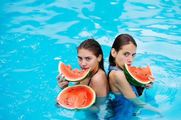 Giovani donne o ragazze sexy con bel viso e capelli bagnati che nuotano in piscina con acqua blu che mangiano anguria rossa soleggiata giornata estiva all'aperto