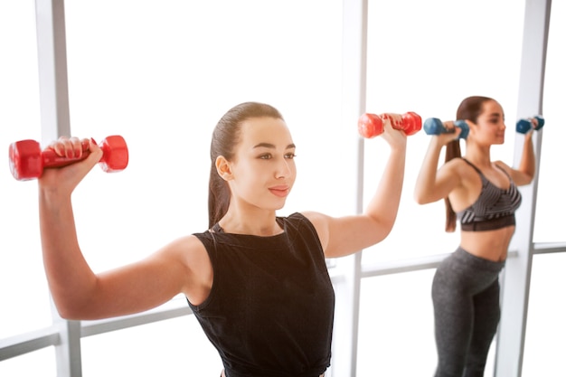 Giovani donne piacevoli e sicure che si esercitano con i dumbbells alla finestra. La modella asiatica sta davanti e guarda avanti. La giovane donna europea è dietro e anche lei si allena