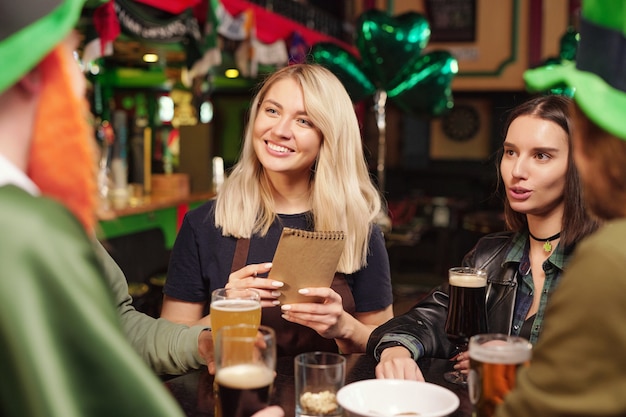Giovani donne sedute al tavolo che parlano con gli amici e bevono birra durante la celebrazione del giorno di San Patrizio al bar
