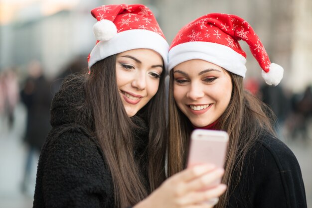 Giovani donne sorridenti in cappello di Natale facendo uso di uno smartphone insieme