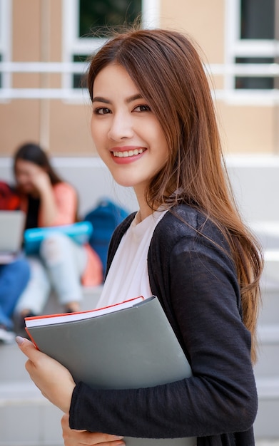 Giovani e belle ragazze studentesse di college asiatiche che tengono libri, posano davanti alla telecamera con un gruppo di amici sfocano sullo sfondo contro l'edificio scolastico. Apprendimento e amicizia del concetto di amico intimo degli adolescenti.