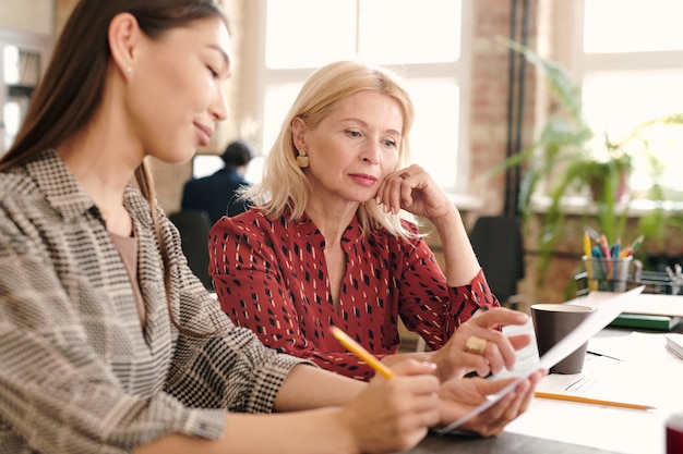 Giovani e mature designer donne serie che guardano uno schizzo su carta mentre uno di loro lo indica durante la discussione dei dettagli durante la riunione