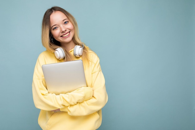 Giovani europei sorridenti studente donna che indossa felpa gialla tenendo il notebook su sfondo blu isolato con spazio libero. concetto di educazione.