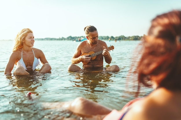 Giovani felici che si divertono insieme in spiaggia. Sono seduti nell'acqua e l'uomo suona l'ukulele e canta. Tramonto sull'acqua.