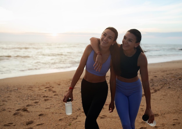 Giovani gemelle europee felici in abiti sportivi che abbracciano il riposo dopo l'allenamento con bottiglie d'acqua