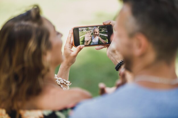 Giovani genitori sorridenti che fanno una videochiamata con la loro figlia carina su uno smartphone mentre si godono una giornata estiva nel parco.