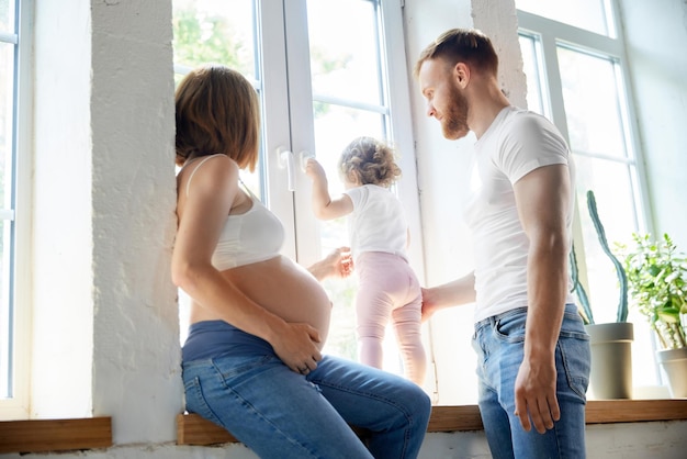 Giovani genitori uomo e donna incinta che giocano con la figlia della bambina a casa guardando fuori dalla finestra Concetto di gravidanza famiglia amore rapporto genitorialità e infanzia