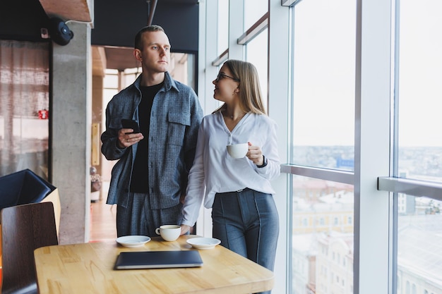 Giovani professionisti intelligenti in abiti casual che controllano i dati al telefono e prendono appunti stando in piedi a un tavolo in un ufficio moderno vicino a una grande finestra all'ultimo piano