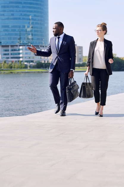 Giovani professionisti interculturali in giacca e cravatta che si spostano lungo il marciapiede lungo il fiume e discutono dell'area mentre tornano dal lavoro