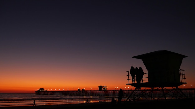 Giovani ragazze adolescenti sagome vicino alla torre del bagnino, amici sulla spiaggia dell'oceano pacifico, tramonto al tramonto a Oceanside, California USA. Adolescenti irriconoscibili, persone e cielo viola viola sfumato crepuscolare.