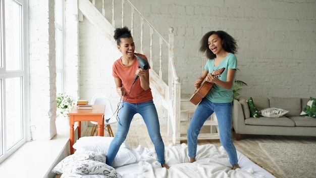 Giovani ragazze divertenti di razza mista ballano cantando con l'asciugacapelli e suonando la chitarra acustica su un letto