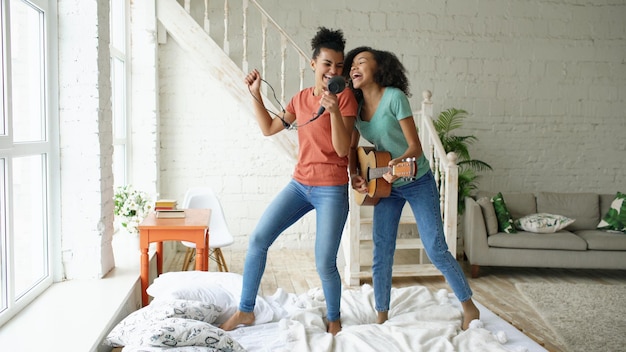 Giovani ragazze divertenti di razza mista ballano cantando con l'asciugacapelli e suonando la chitarra acustica su un letto